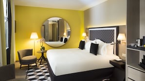 Ein Hotelzimmer mit einem großen Bett, zwei Nachttischen, einem runden Spiegel, einem Sessel und einem kleinen Tisch mit Lampe und Blumen.