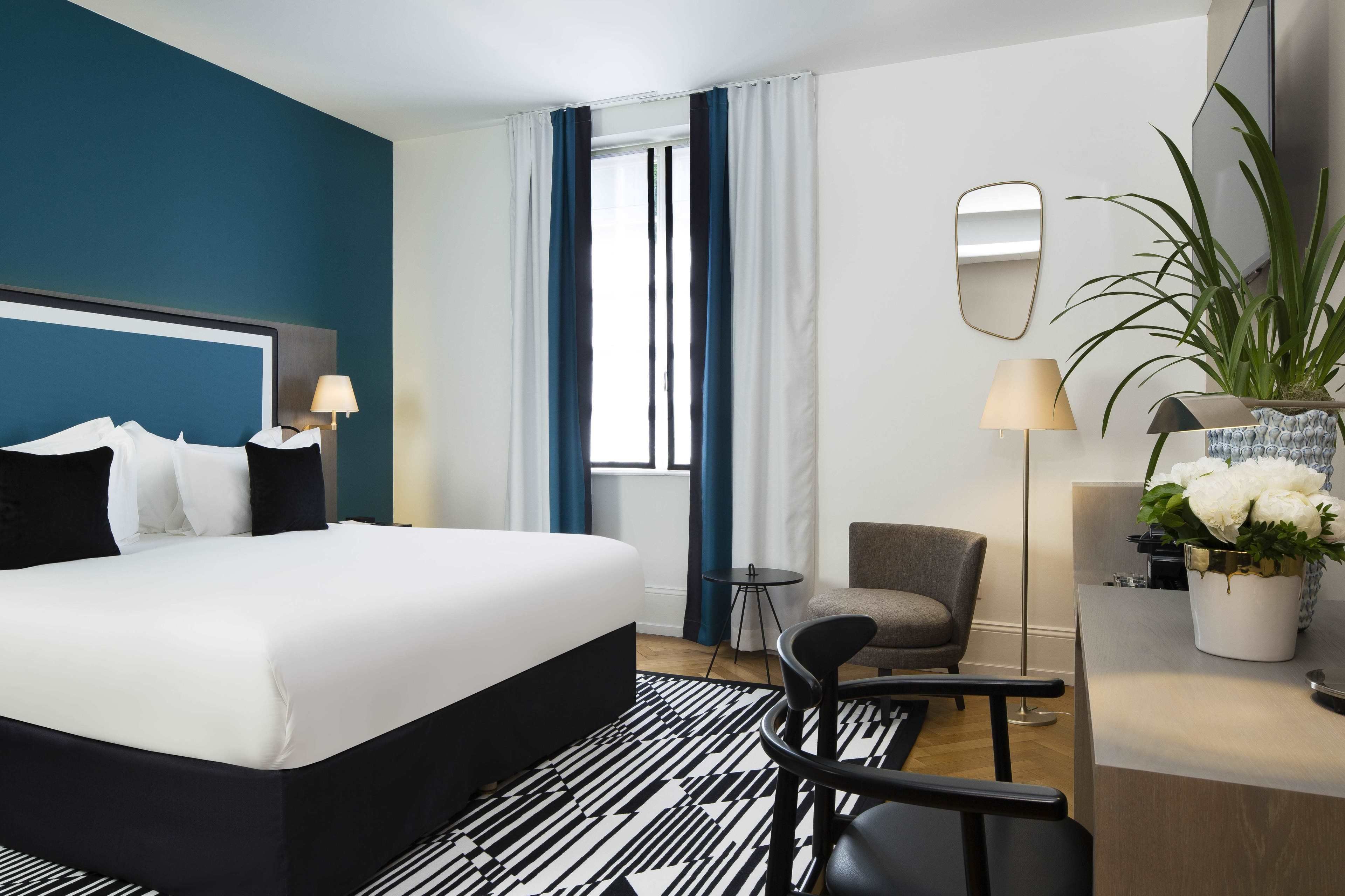A hotel room with a large bed, a desk with a lamp, a chair, a small table, a potted plant, and a window with curtains.