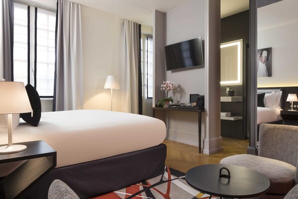 A hotel room with a large bed, a desk with a lamp, a small round table, a sofa, and a TV mounted on the wall. - Boutique Hôtel des XV (Strasbourg)