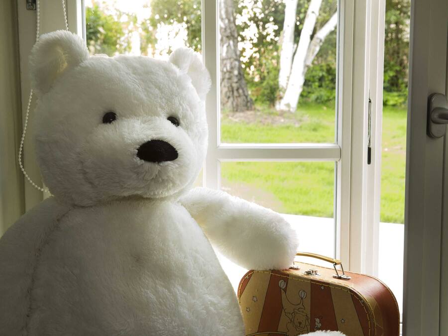Un ours en peluche blanc est posé à côté d’une valise ancienne, près d’une fenêtre.