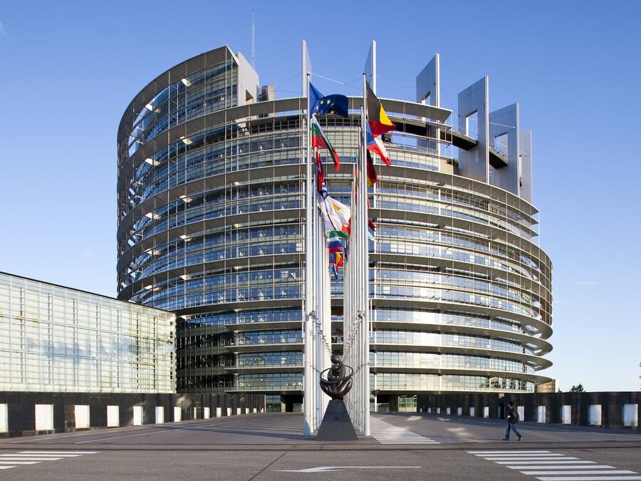 Un edificio moderno y curvo con varias banderas, incluyendo la bandera de la Unión Europea, y un paso de peatones al frente.