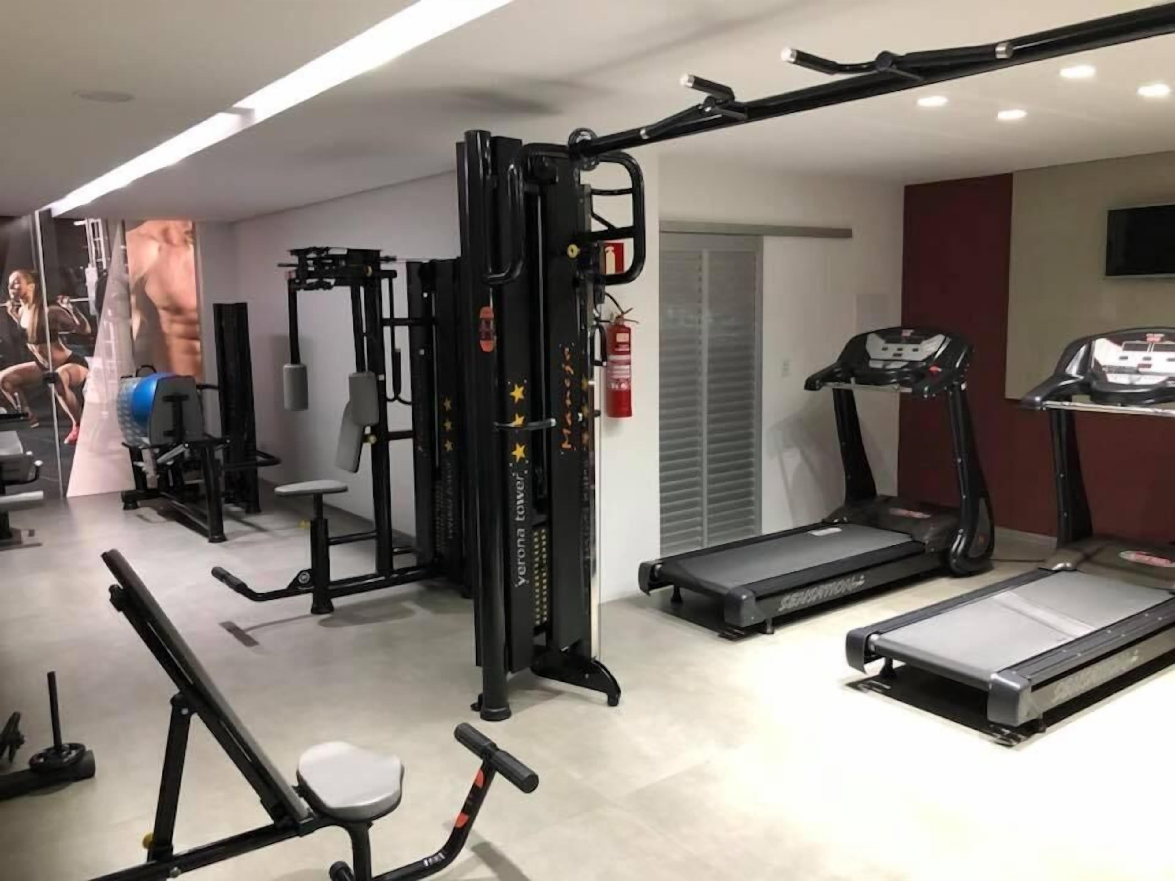 Uma academia com diversos equipamentos de exercício, incluindo um banco de supino, um suporte para agachamento e esteiras.