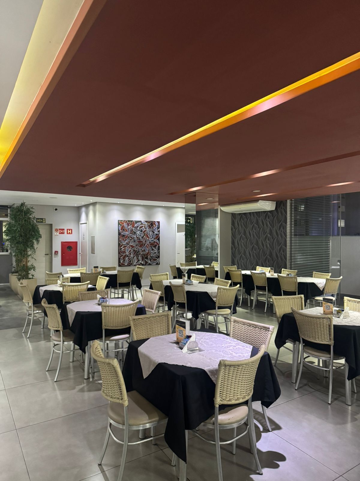 Un comedor moderno con mesas redondas y sillas de mimbre, un detector de incendios y un cuadro abstracto grande en la pared.