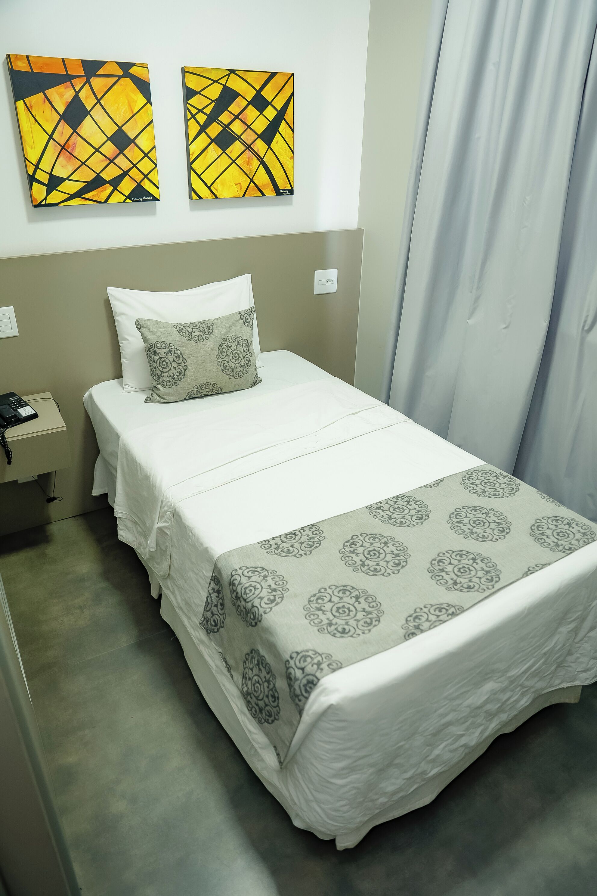 A neatly made bed with a patterned bedspread, a bedside table with a phone, and two abstract paintings on the wall.
