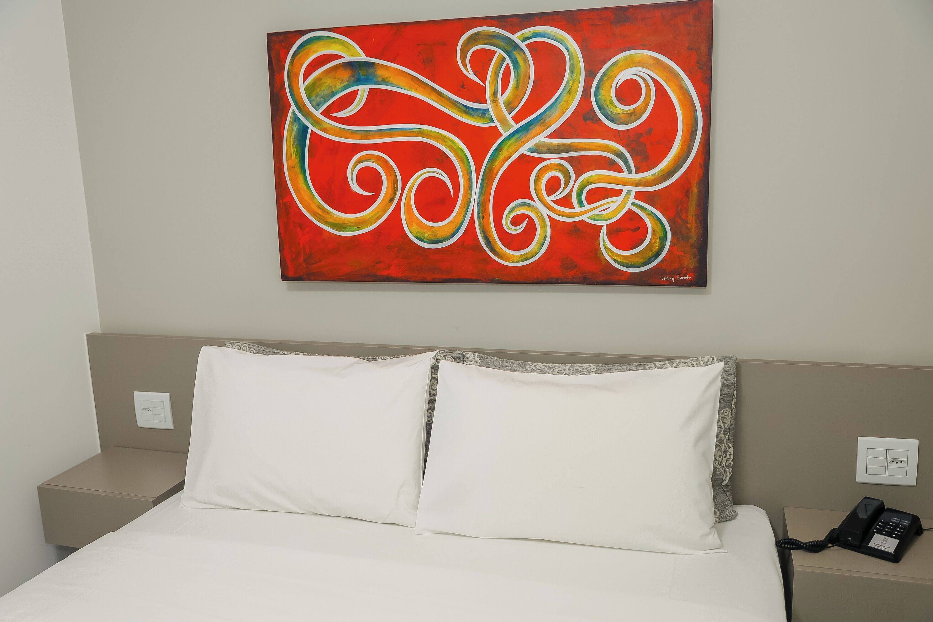 Habitación de hotel con cama, almohadas blancas, un cuadro abstracto rojo en la pared, teléfono en la mesita de noche y toma de corriente.