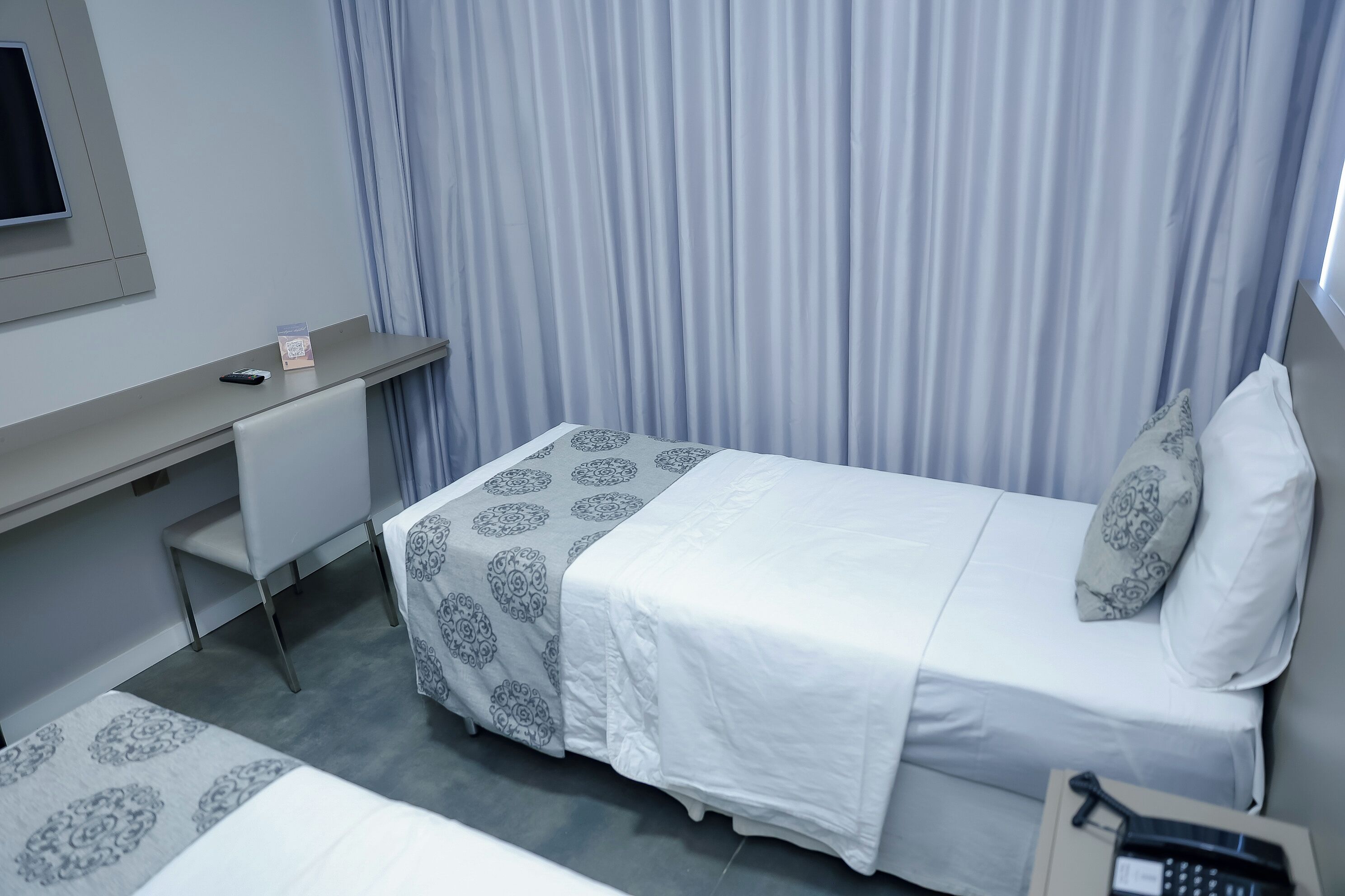 A hotel room with a bed, a desk, and a chair. There is a telephone on the desk and a package on the floor.