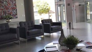 Un moderno vestíbulo con tres sofás oscuros, una mesa redonda con revistas y una planta, y un cuadro abstracto grande en la pared.