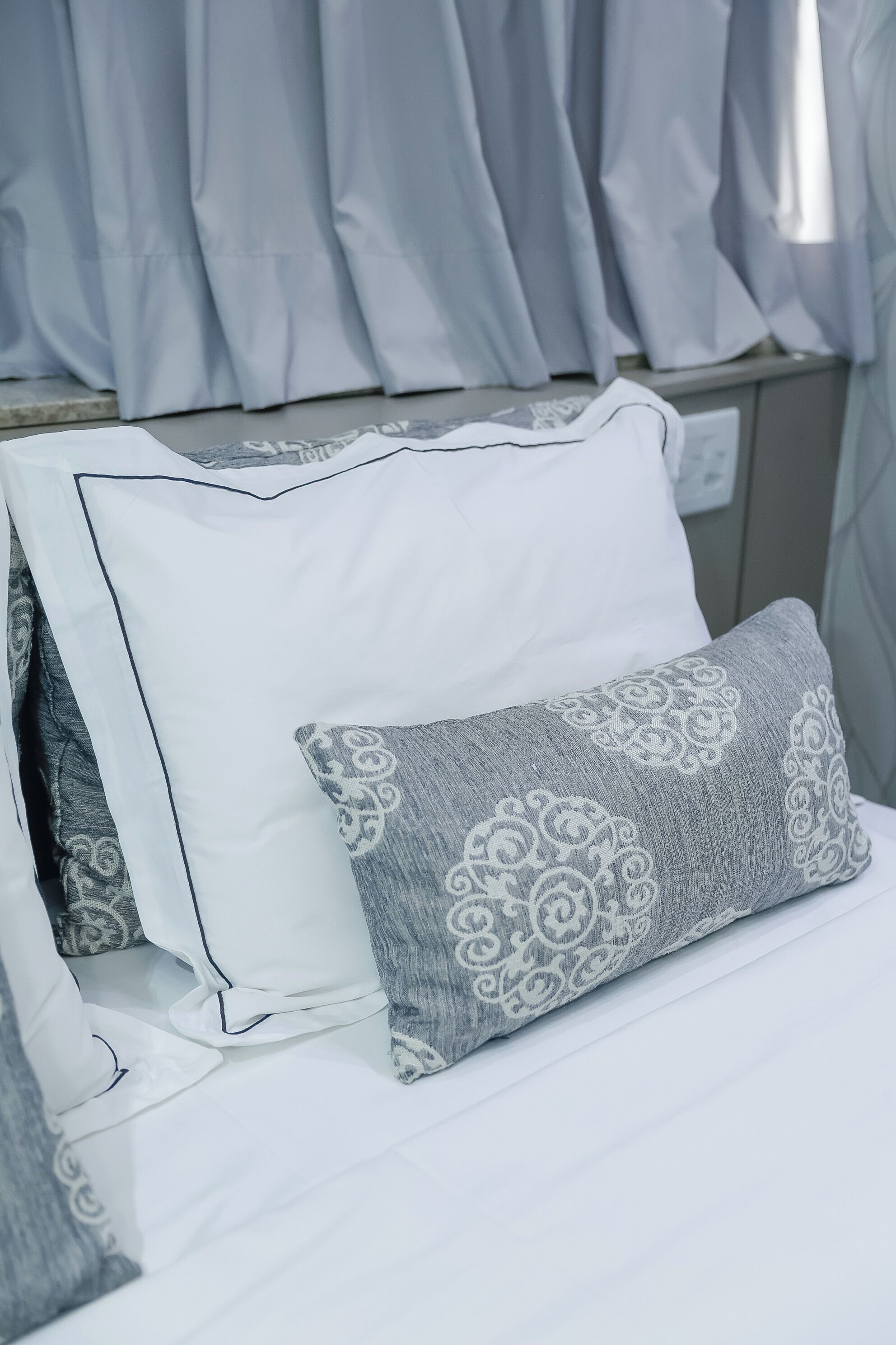Uma cama com almofadas brancas e cinzentas, cabeceira branca e cortinas claras.