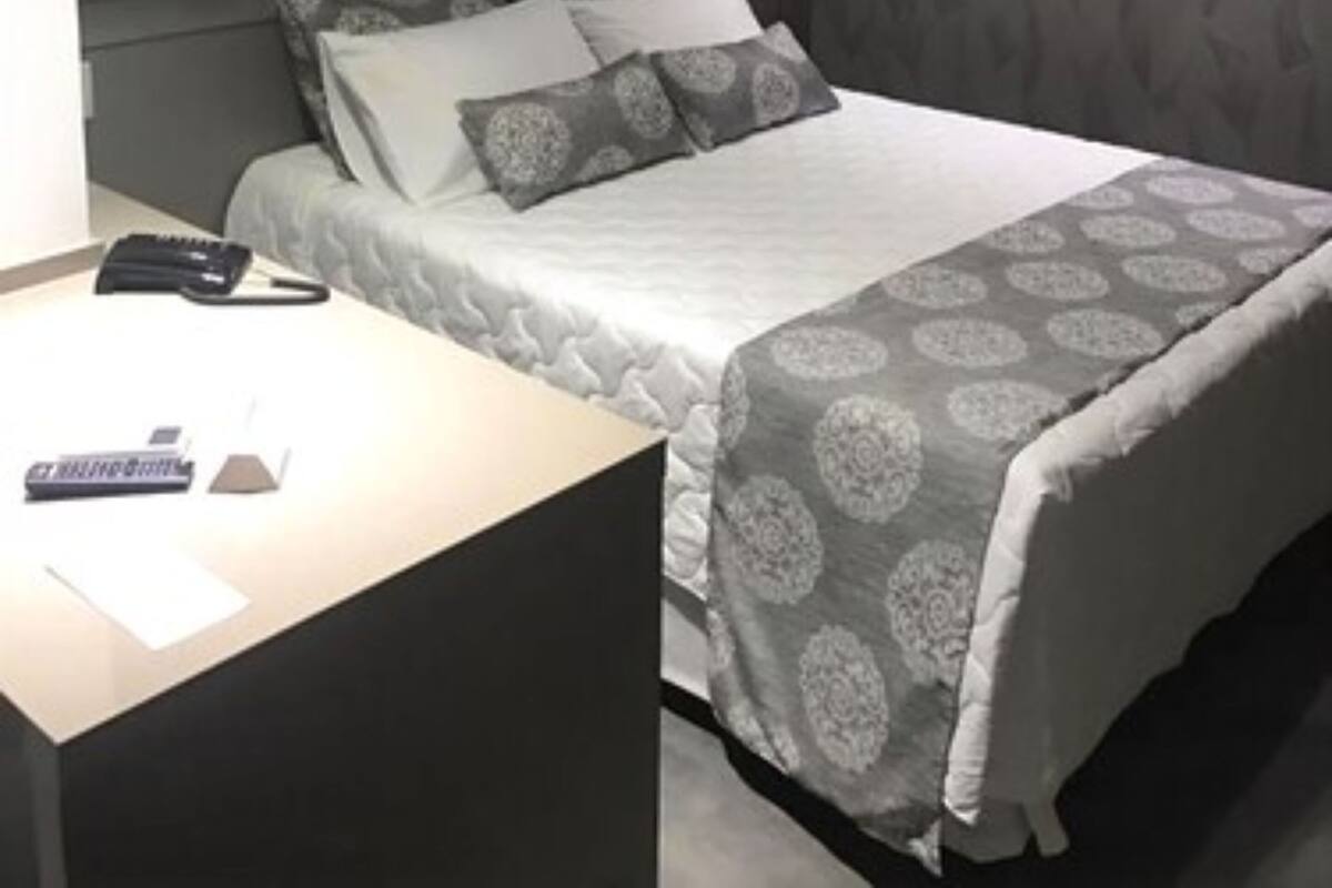 A hotel room with a bed, a bedside table, a desk, and an air conditioning unit.
