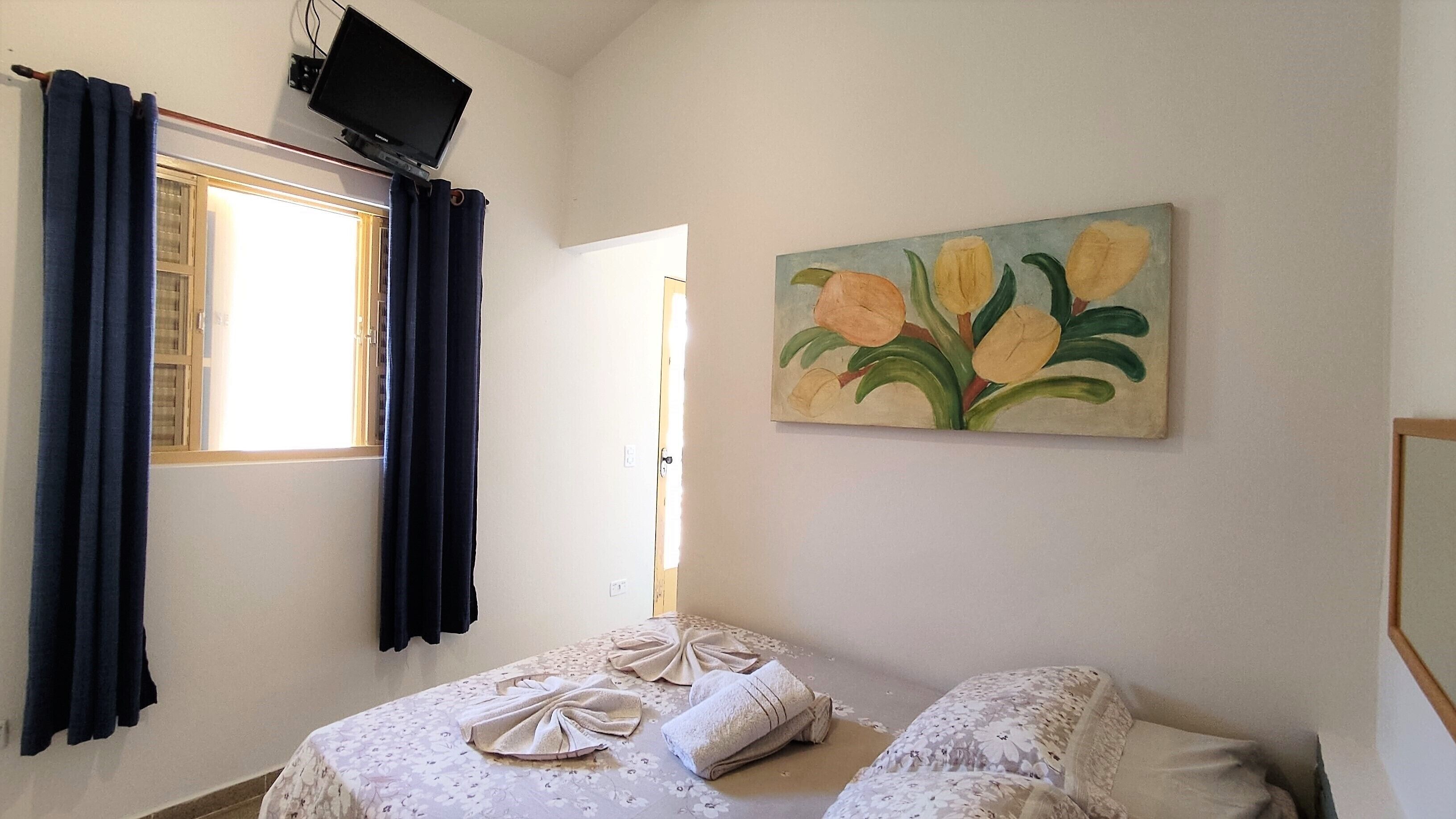 A bedroom with a bed, a painting of flowers on the wall, and a window with curtains.