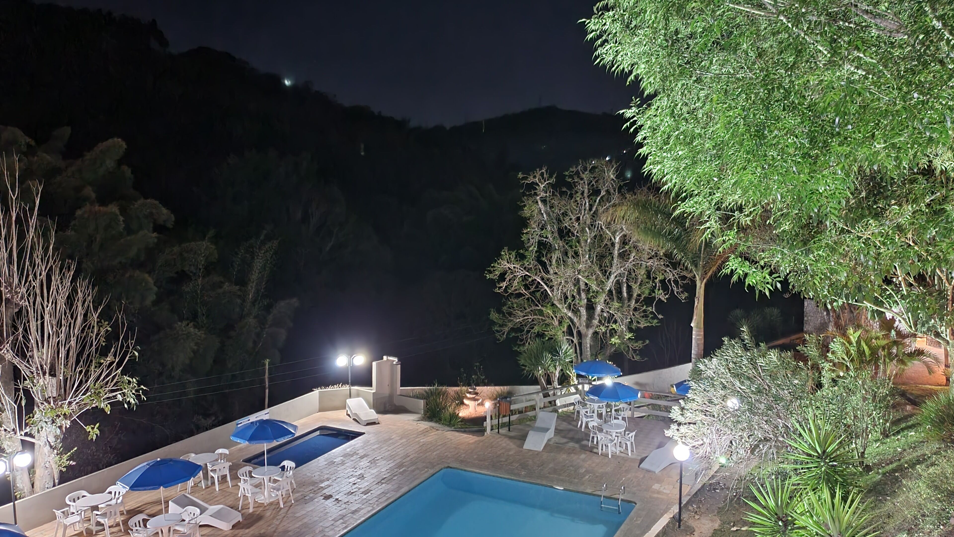 A nighttime pool area with lounge chairs and umbrellas, surrounded by trees and hills.