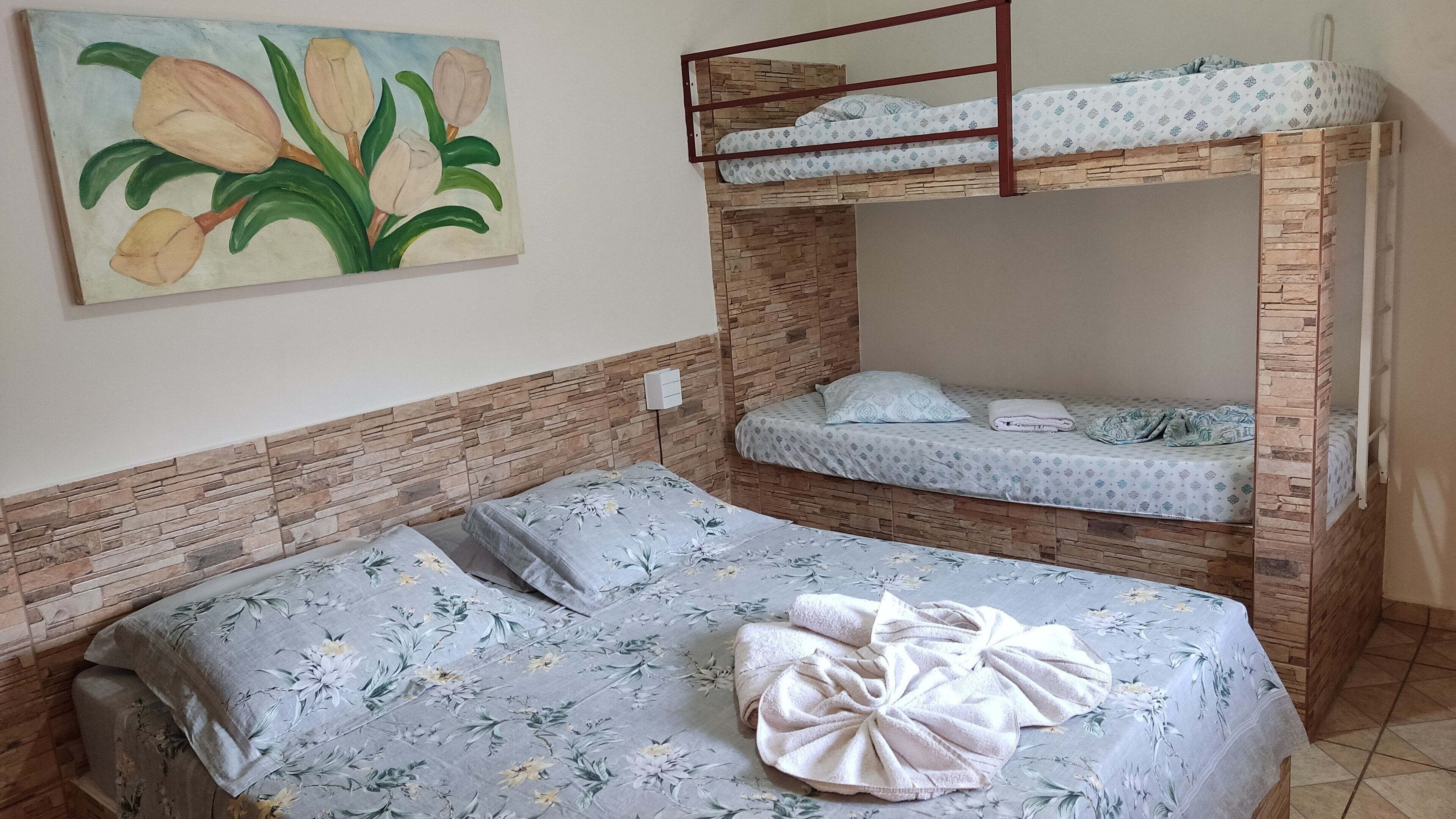 A room with a bunk bed, a single bed, floral bedding, a painting of flowers, and a stone-patterned wall.