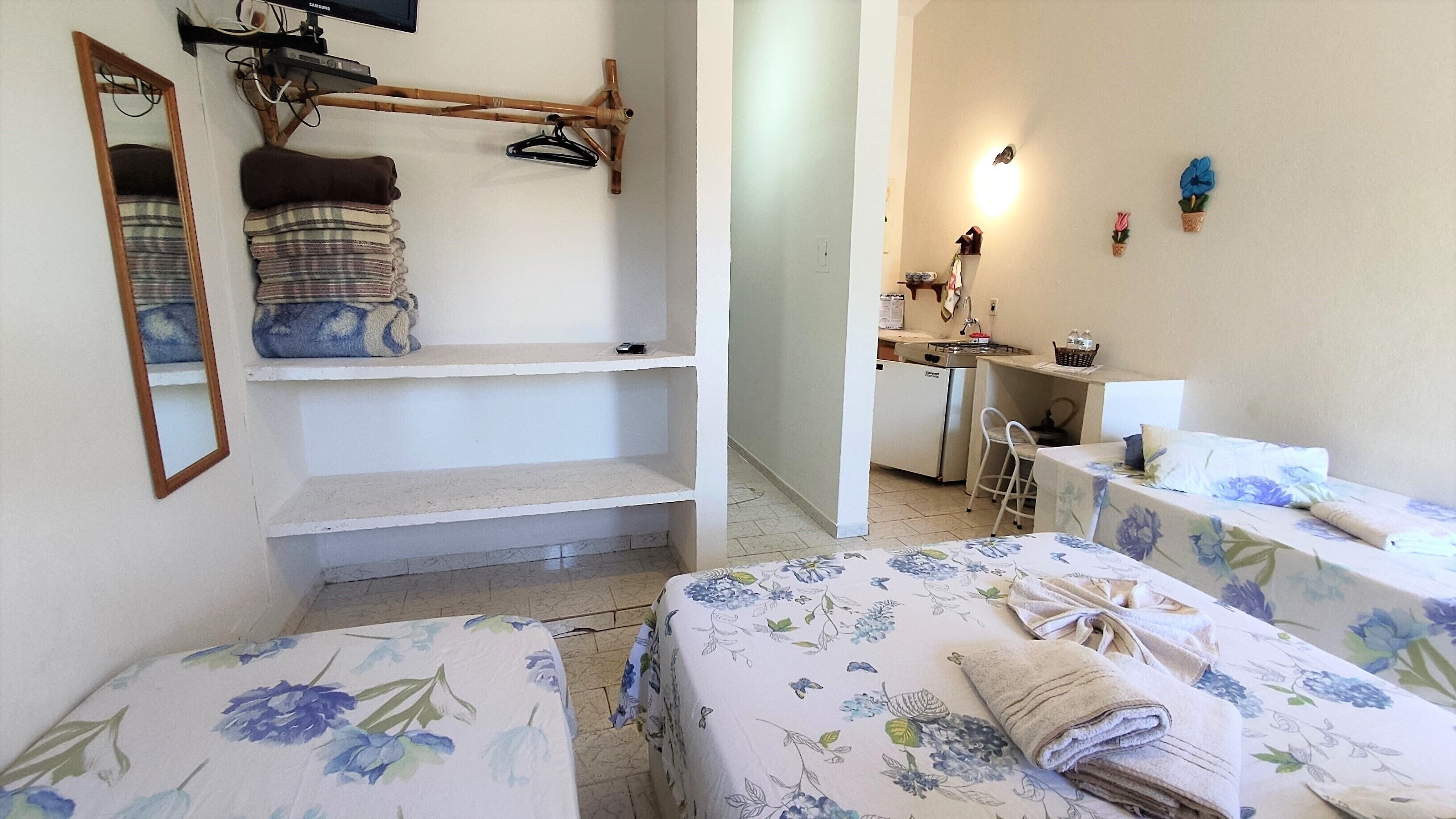 A bedroom with two beds, a small kitchenette, a mirror, and a shelf with folded towels.