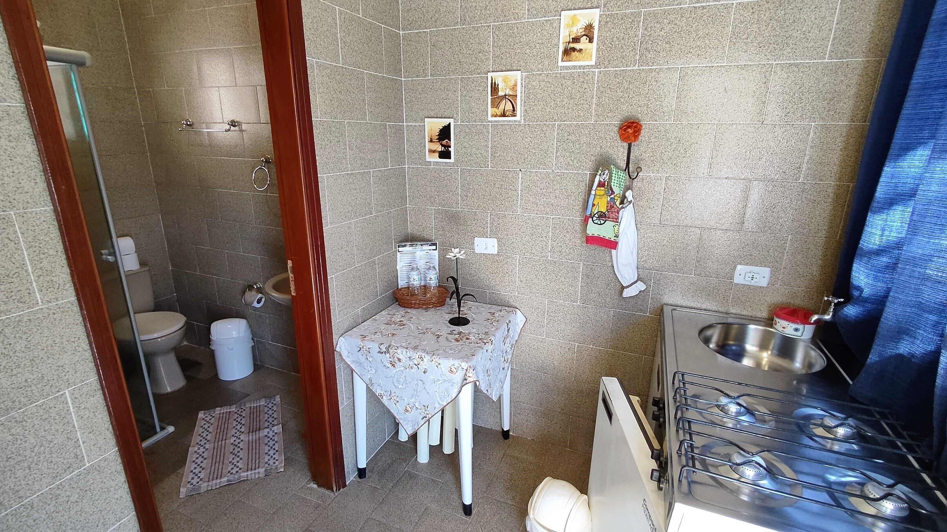 A compact bathroom with a toilet, sink, and shower. There is a small table with a floral tablecloth and a basket of toiletries.