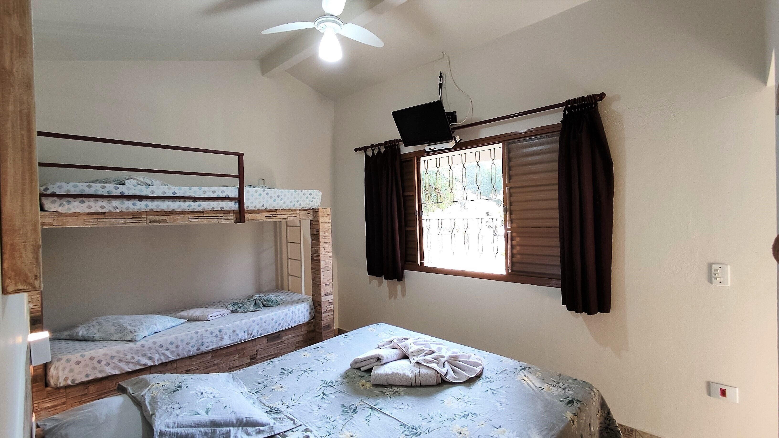 A dormitory room with bunk beds, a single bed, a ceiling fan, a window with curtains, and a wall socket.