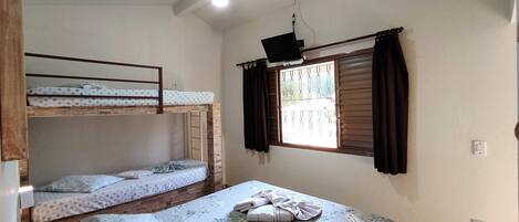 A dormitory room with bunk beds, a single bed, a ceiling fan, a window with curtains, and a wall socket.
