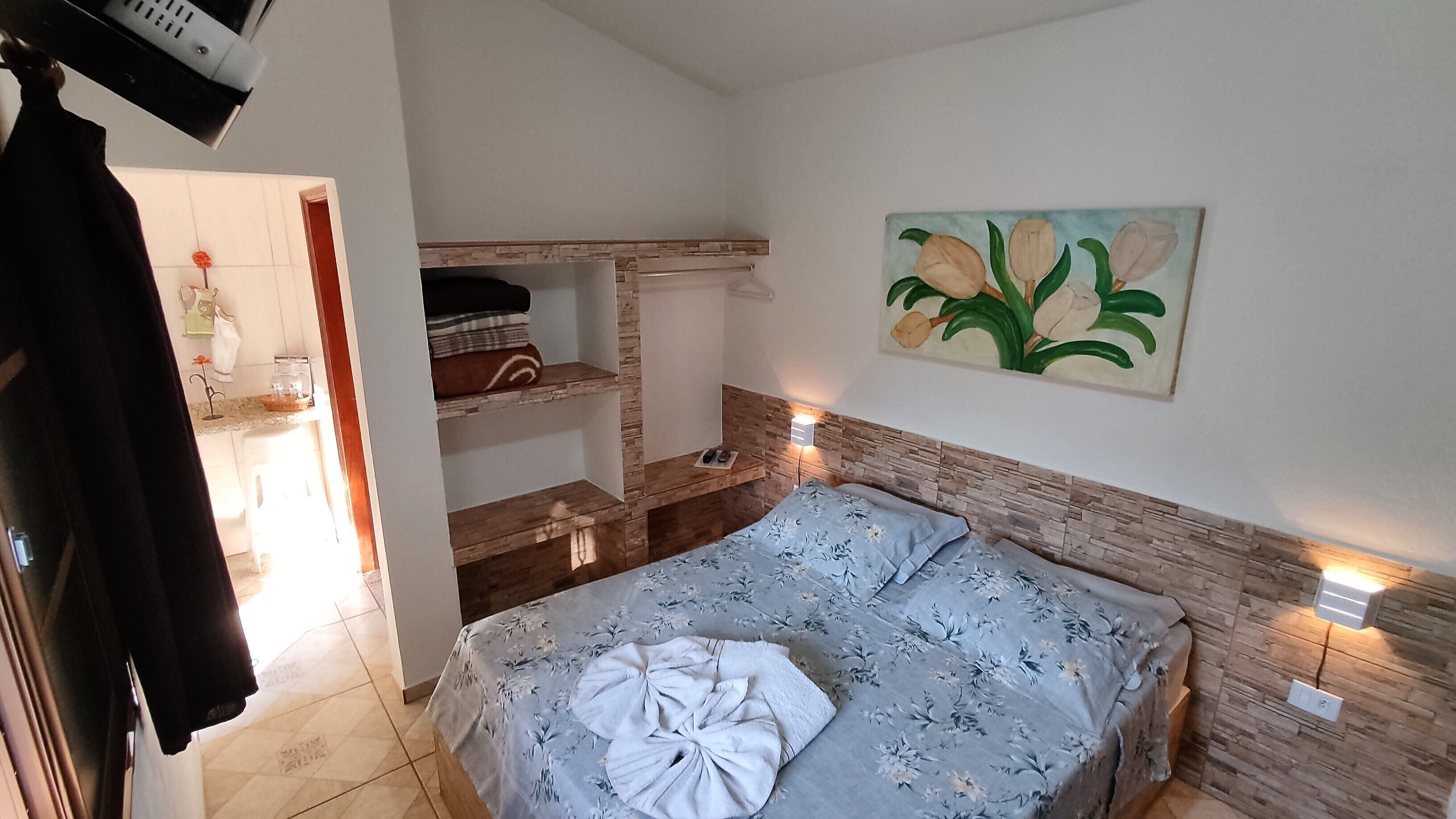 A bedroom with a bed, a painting, a shelf with towels, and a small table with a remote control.