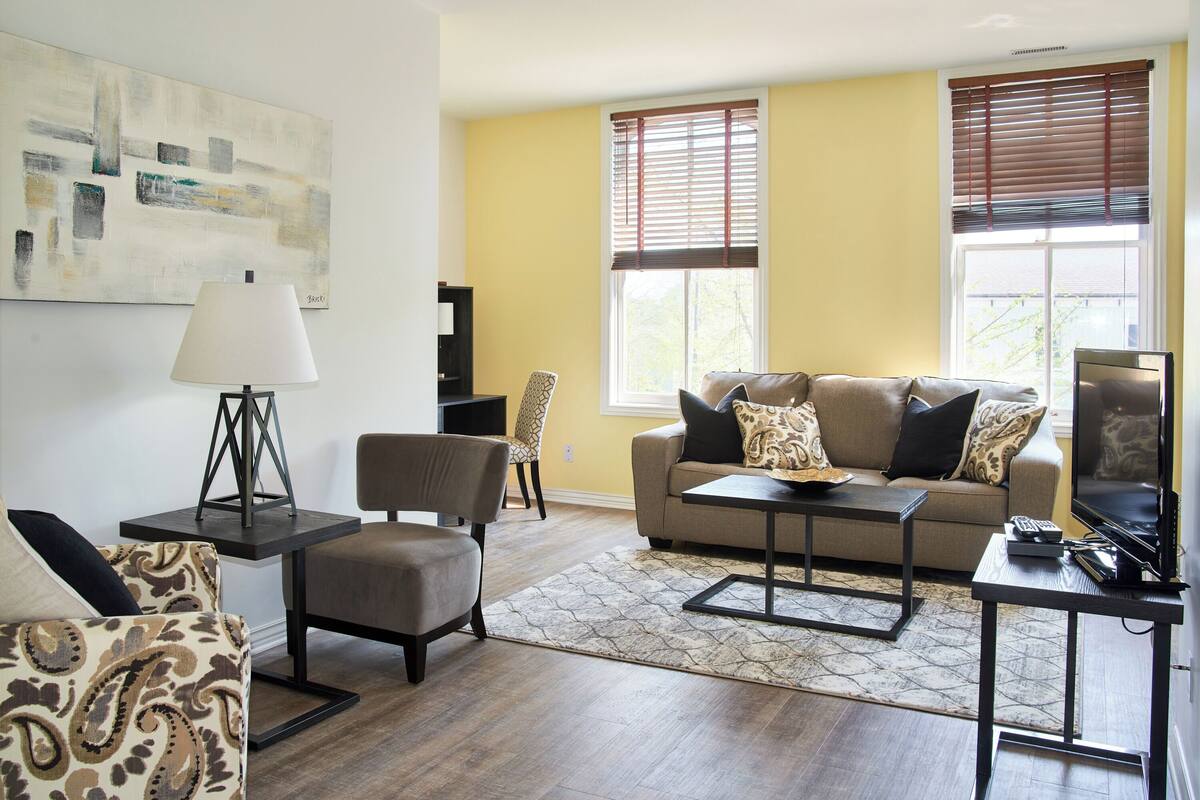 Un salon comprenant un canapé, un fauteuil, une table basse et un téléviseur. Il y a un tableau au mur et une fenêtre avec des stores.