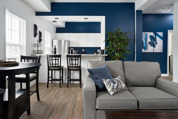 A modern kitchen with white cabinets, a gray sofa with a patterned pillow, and a dining area with a wooden table and chairs.