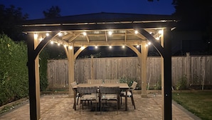 Un'area patio coperta con tavolo e sedie, circondata da un recinto in legno e un vialetto in mattoni.