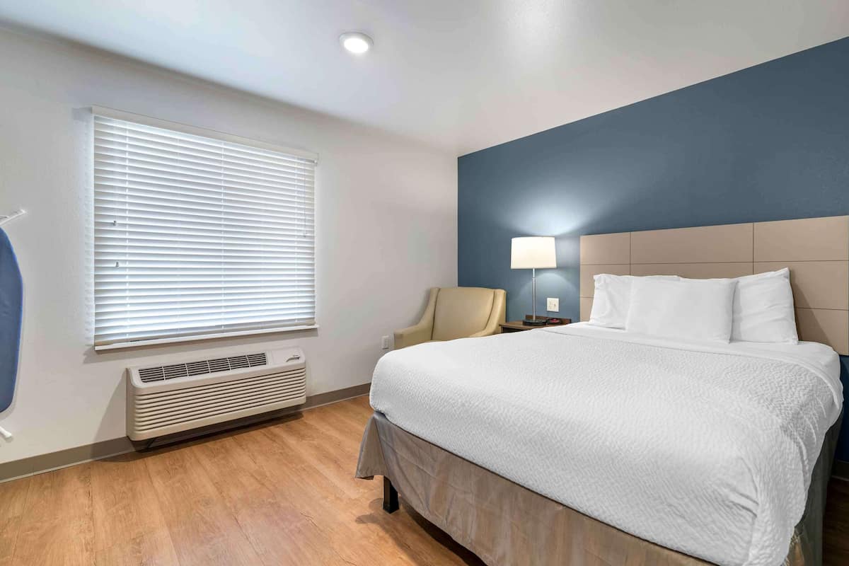 A hotel room with a bed, a chair, a lamp, and a window with blinds.