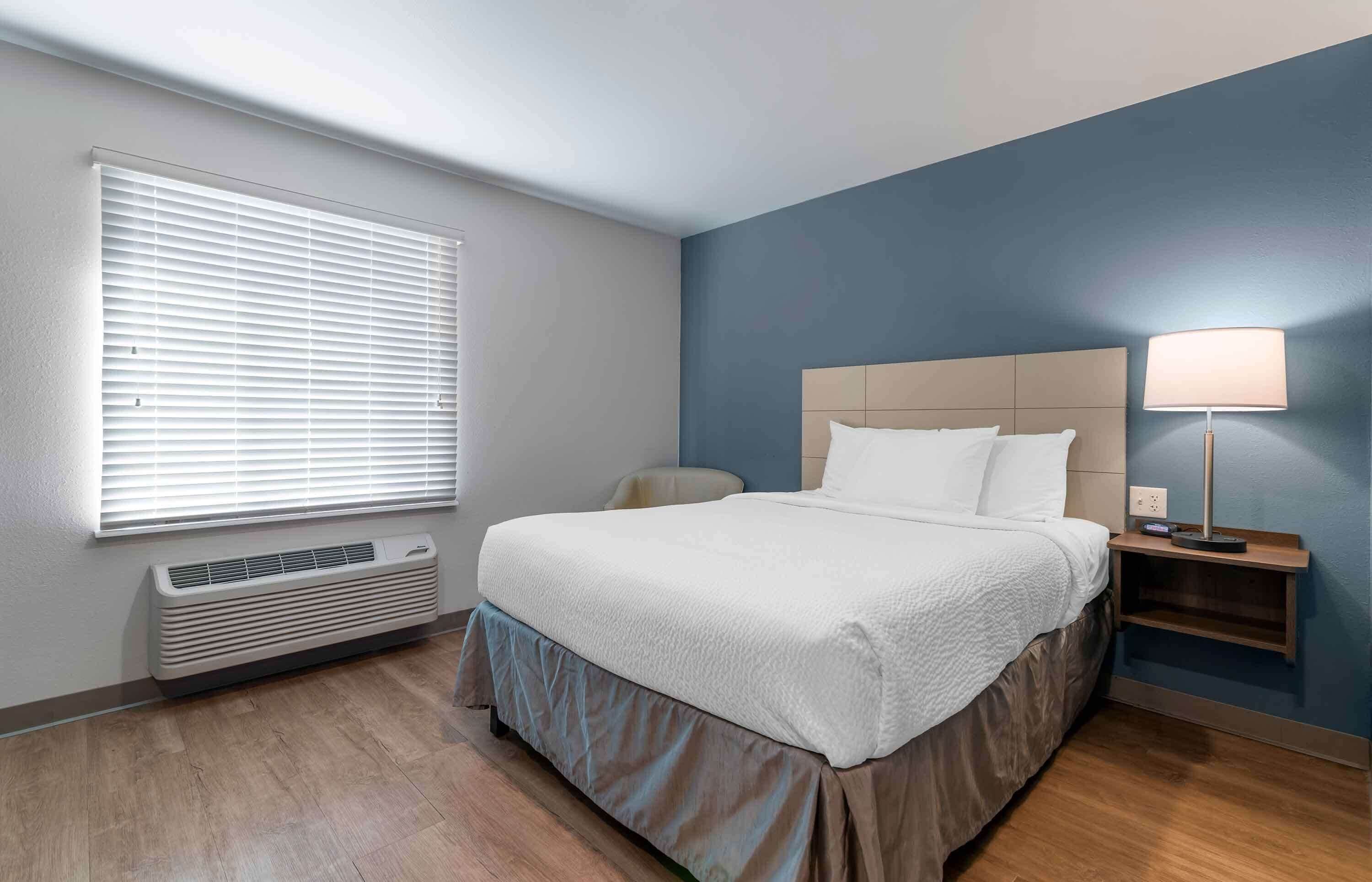 Habitación de hotel con cama, mesita de noche, lámpara, equipo de aire acondicionado y ventana con persianas.