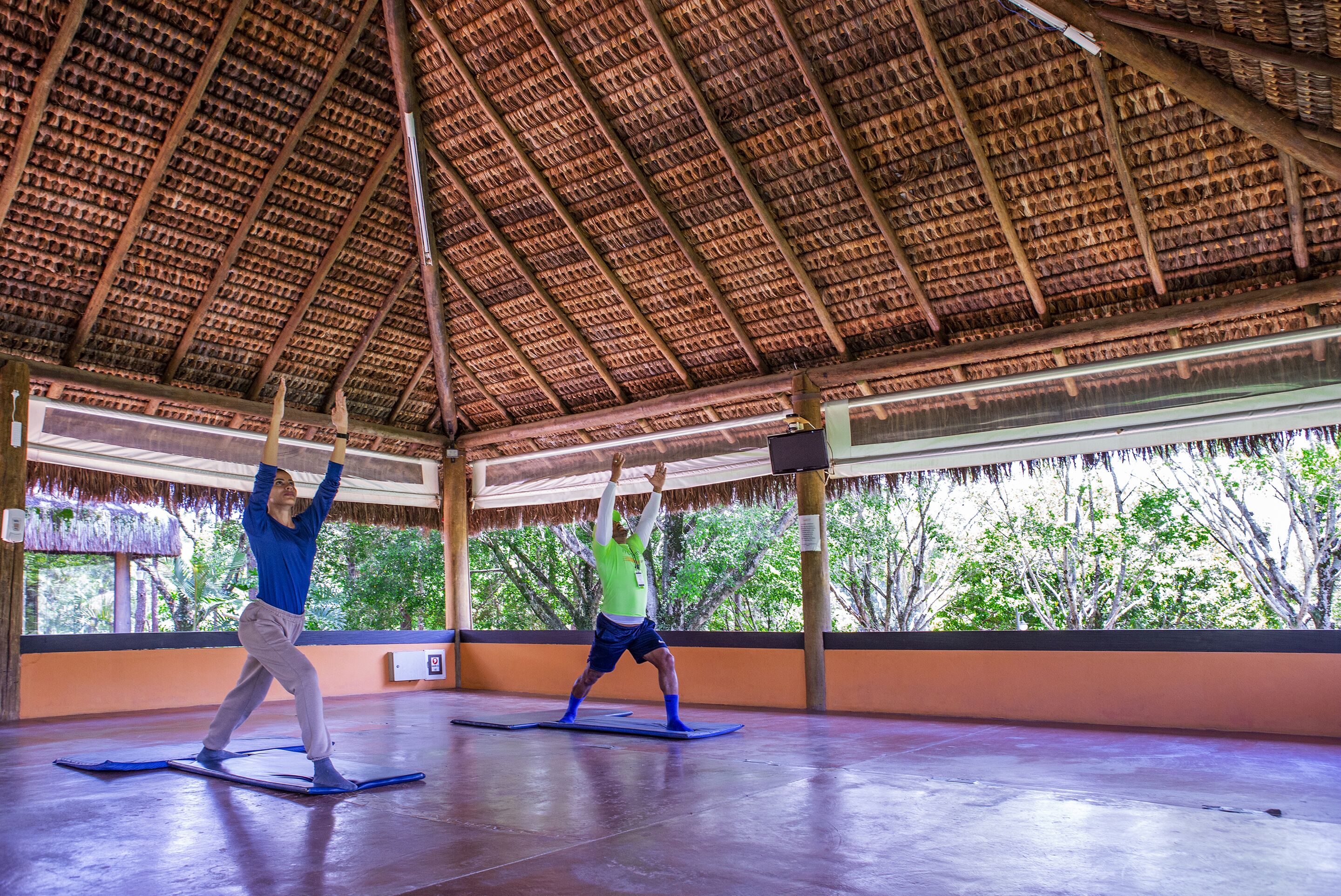 Amplio espacio interior con techo alto y techo de paja, donde dos personas practican yoga sobre colchonetas.