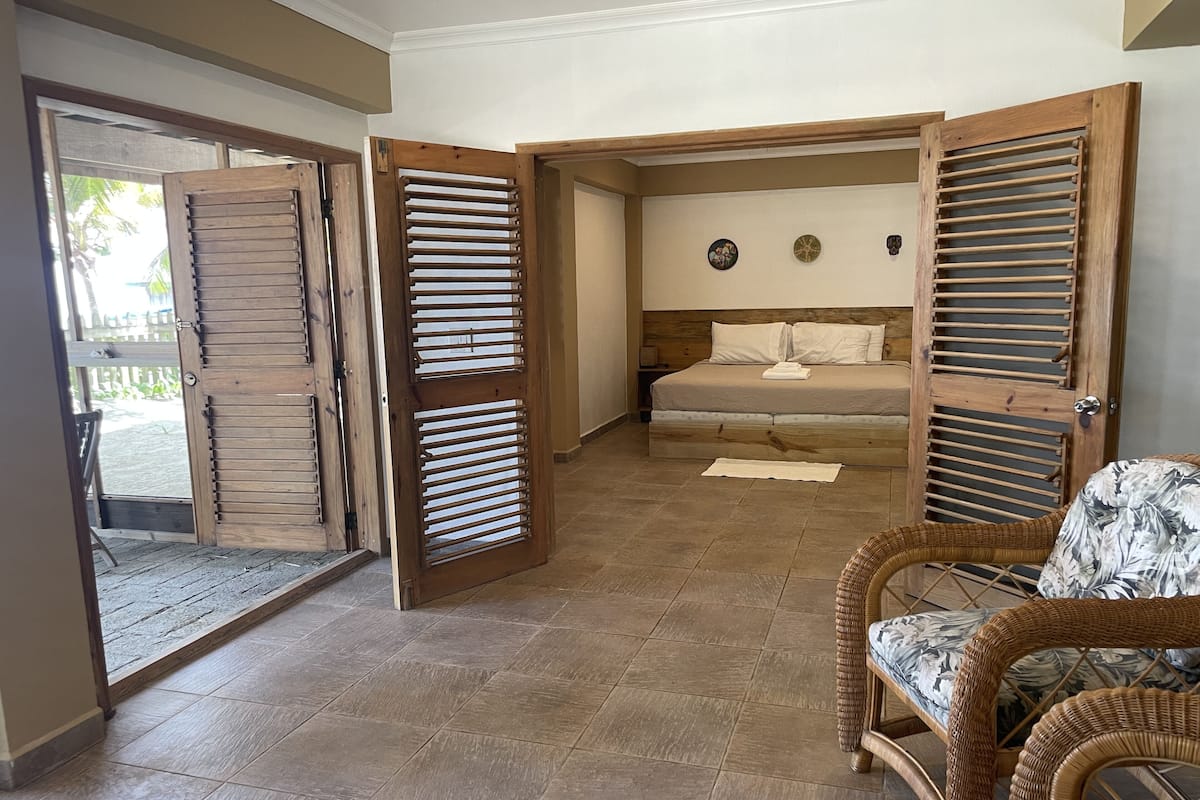 A room with a bed, a wicker chair, and a wooden door with louvered panels.
