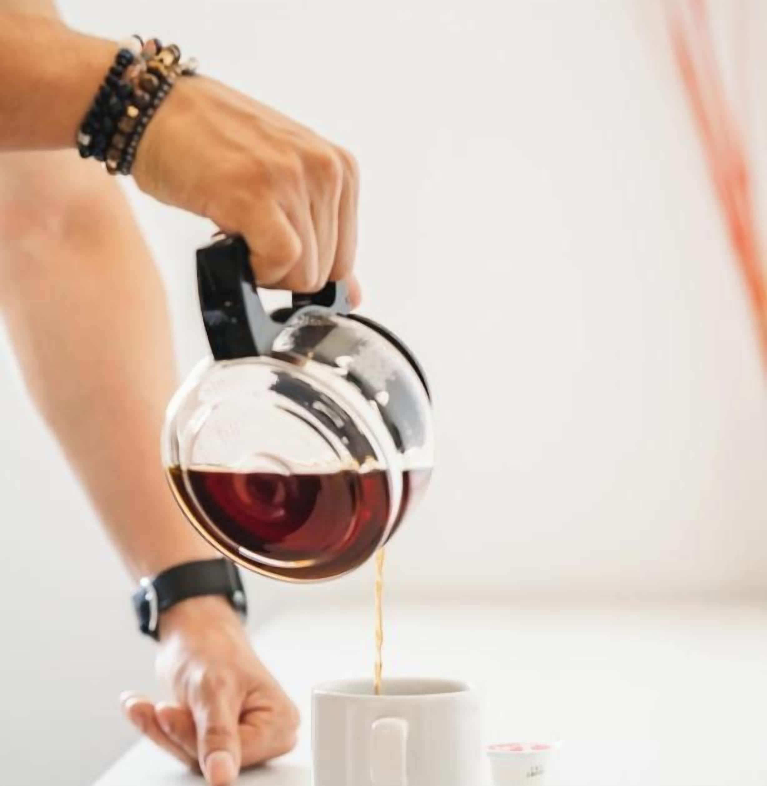 A person pouring a beverage from a metallic kettle into a white mug.