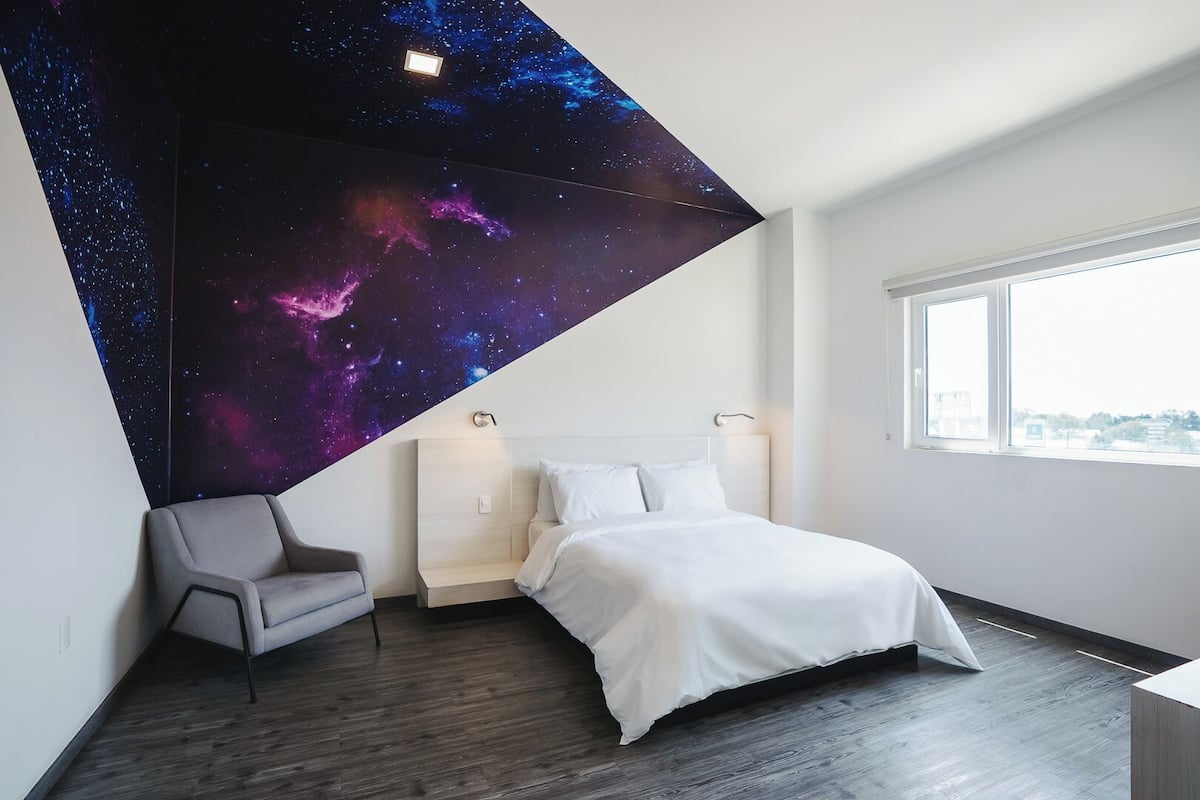 A modern bedroom with a bed, a chair, a nightstand, and a starry sky mural on the ceiling.