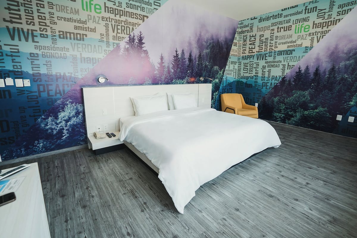 A modern bedroom with a large bed, a nightstand, and a chair. The room features a wooden floor and a wall with a nature-themed mural.