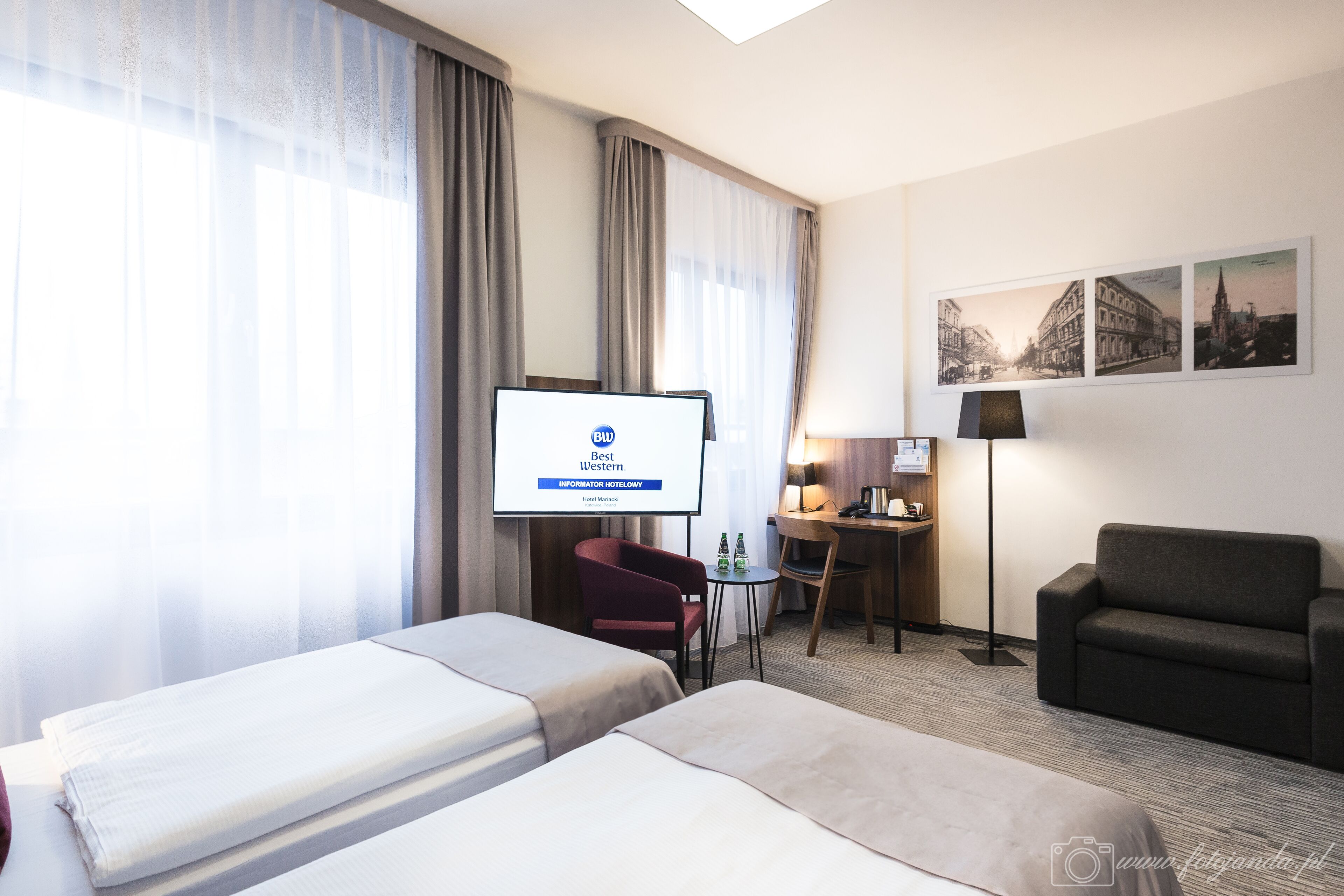 A hotel room with two beds, a desk, a chair, a TV screen, and a lamp.