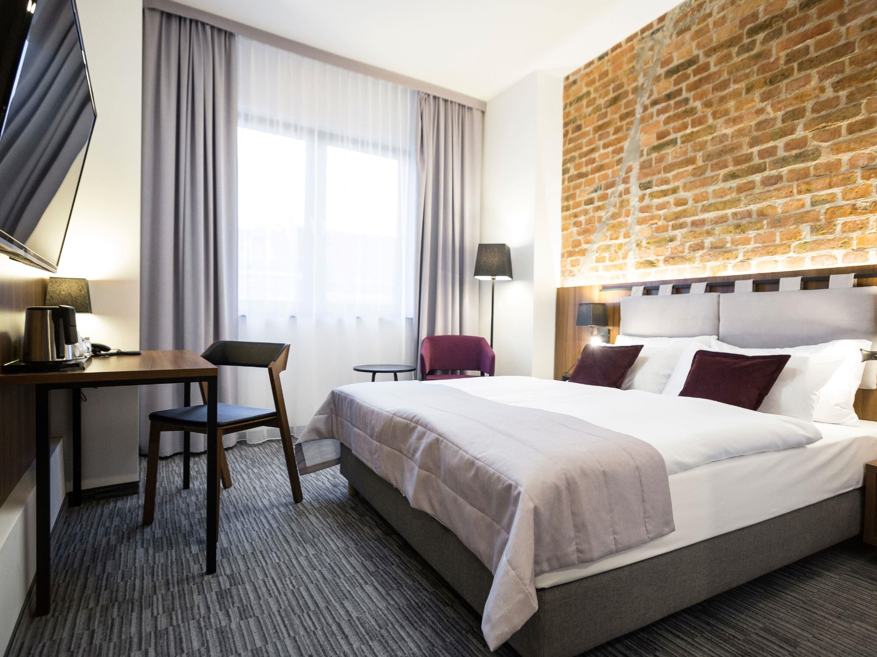 A hotel room with a bed, a desk, a chair, a small table, and a TV.