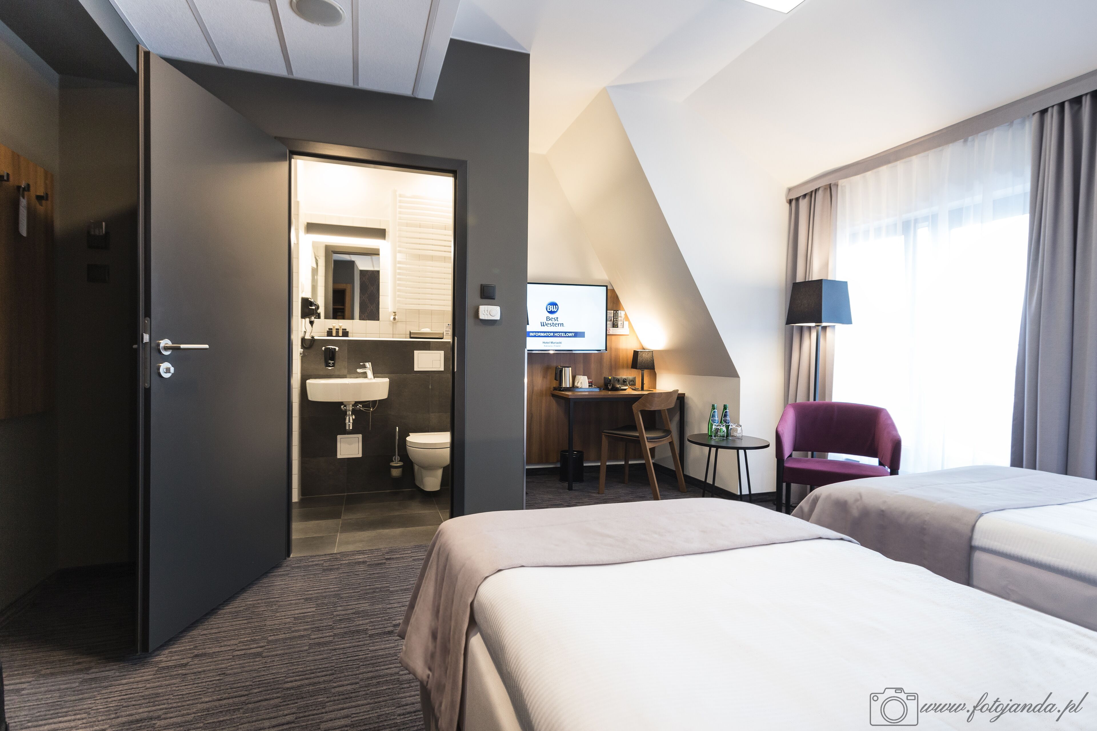 Une chambre d’hôtel avec deux lits, un bureau, une chaise, un lavabo et une salle de bain visible à travers une porte ouverte.