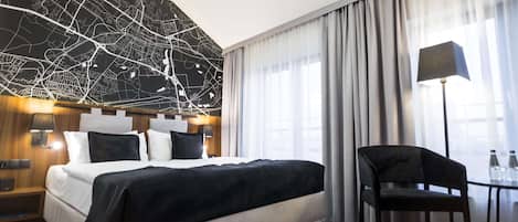 A modern hotel room with a large bed, a desk with a chair, a map wall, and a view of the city through large windows.