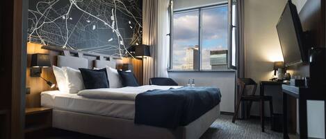 Una habitación de hotel moderna con una cama grande, un escritorio con silla, televisor y vistas a la ciudad por la ventana.