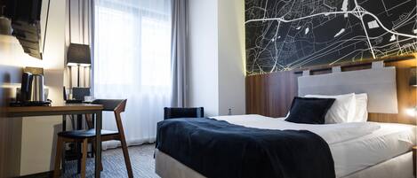 A hotel room with a bed, a desk with a chair, a lamp, a coffee maker, and a large wall map.