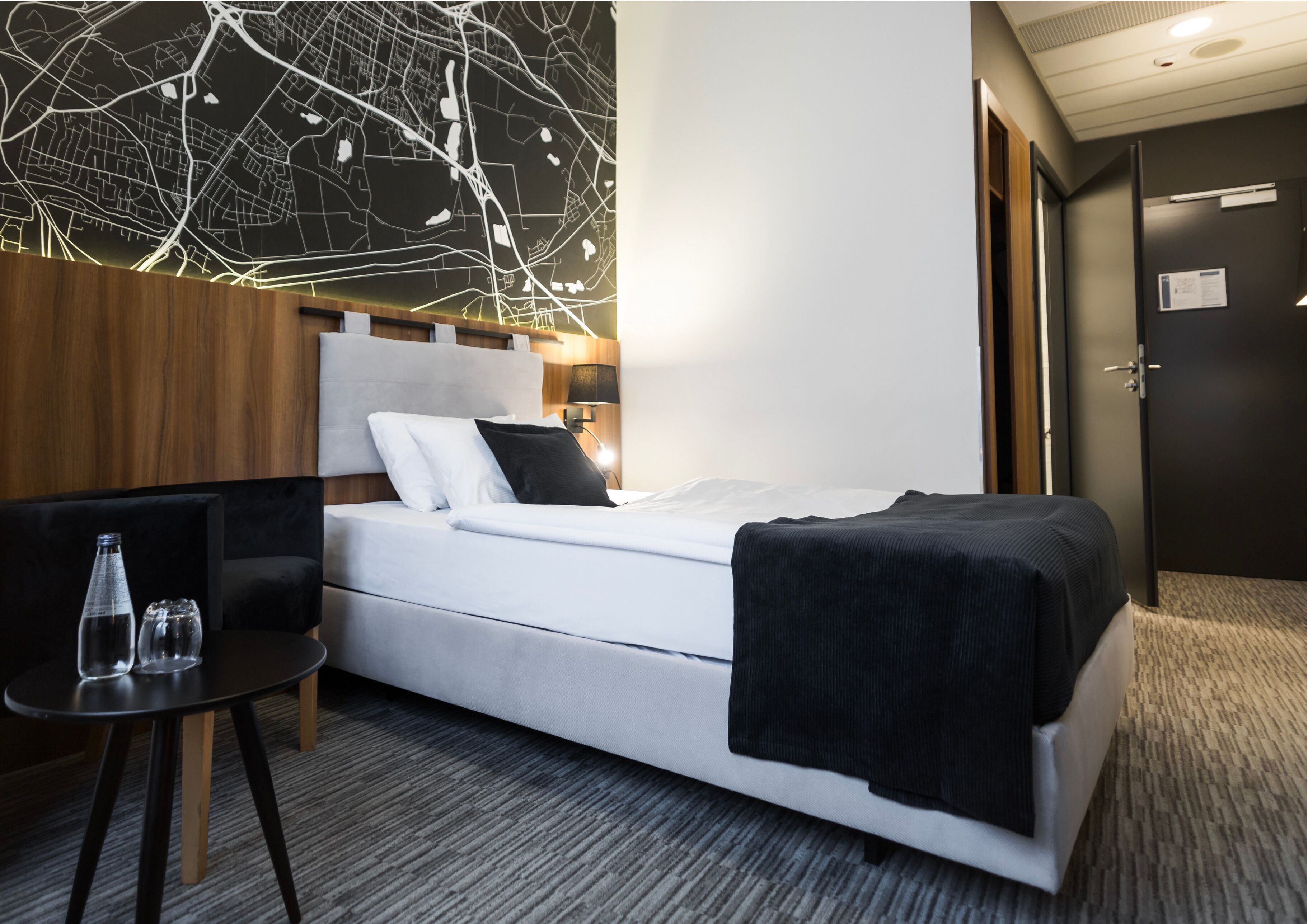Moderni hotellihuone, jossa on siististi pedattu sänky, pieni pöytä laseineen ja seinälle kiinnitetty kartta.