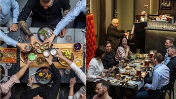 A dining area with multiple tables, some with people seated and others standing. There are various dishes and drinks on the tables.