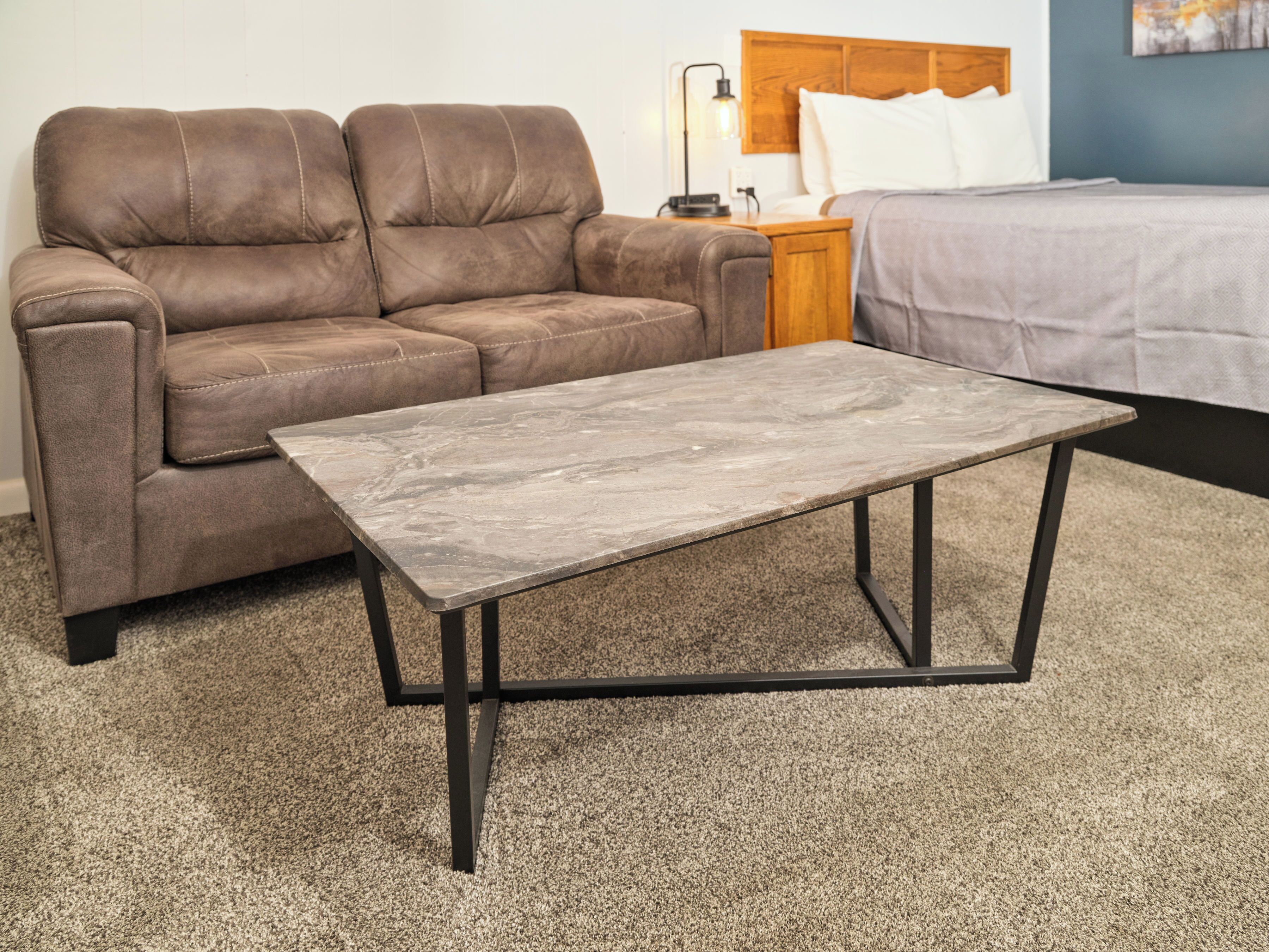 A brown leather sofa, a grey marble coffee table, and a bed with a wooden headboard in a room with a carpeted floor.