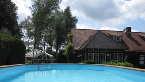 Ein Schwimmbad mit Holzterrasse, umgeben von Bäumen und einem Haus mit Ziegeldach.