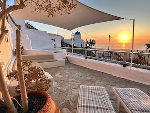 Una terraza con suelo de piedra, un edificio blanco con cúpula azul y vistas al atardecer.