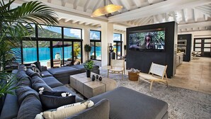 A spacious living area with a large sofa, a coffee table, and a TV. There are plants, a chair, and a view of the ocean. - The BEST Wedding Spot! 2 pools, gym, beachfront & MUCH MORE! (St. Thomas)