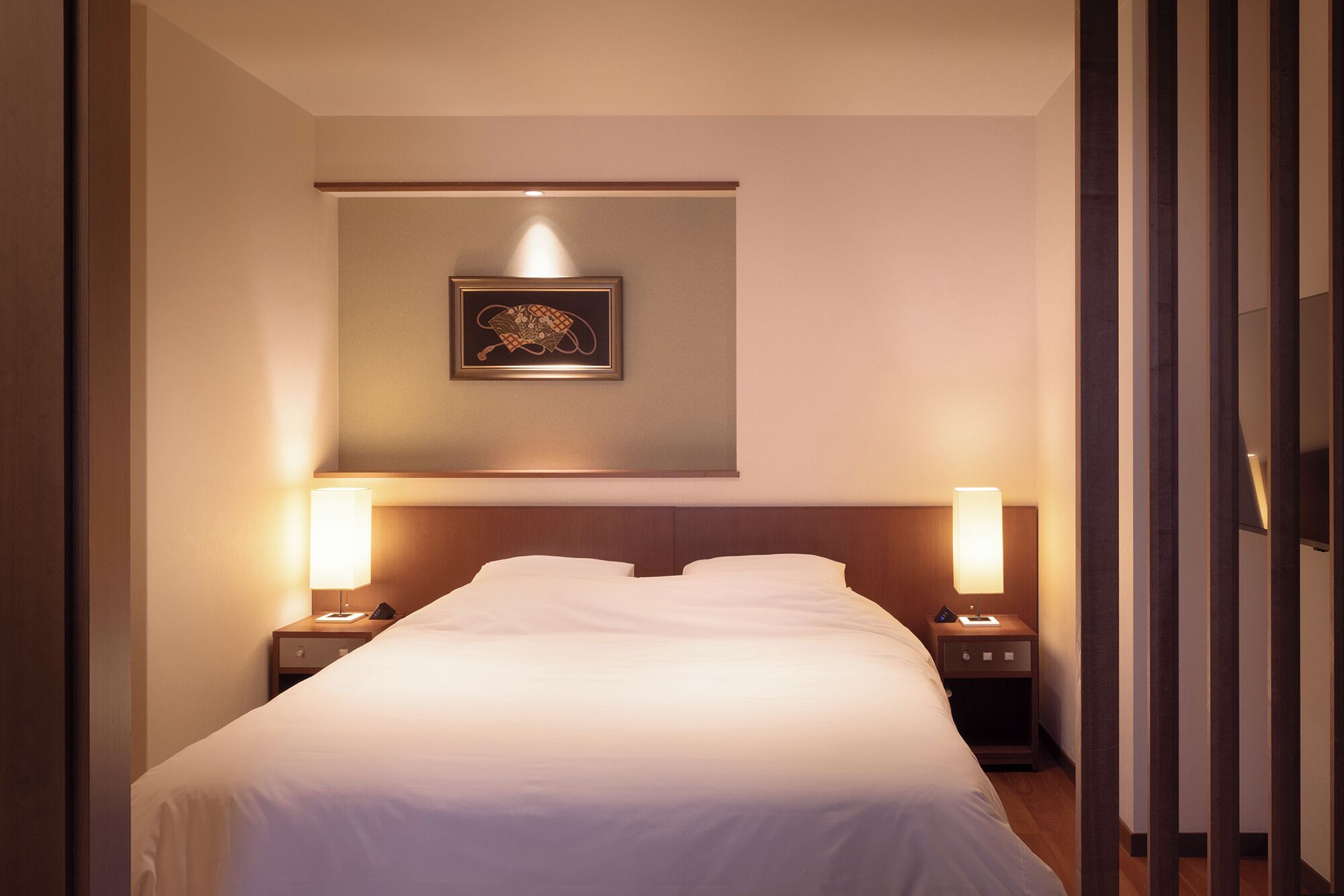 A hotel room with a neatly made bed, two bedside lamps, a wall-mounted telephone, and a framed picture above the bed.