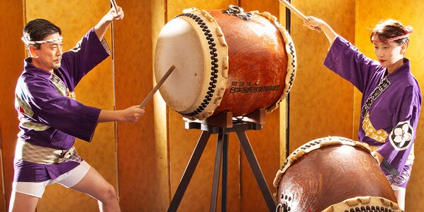 Two individuals playing drums in a room with wooden walls.
