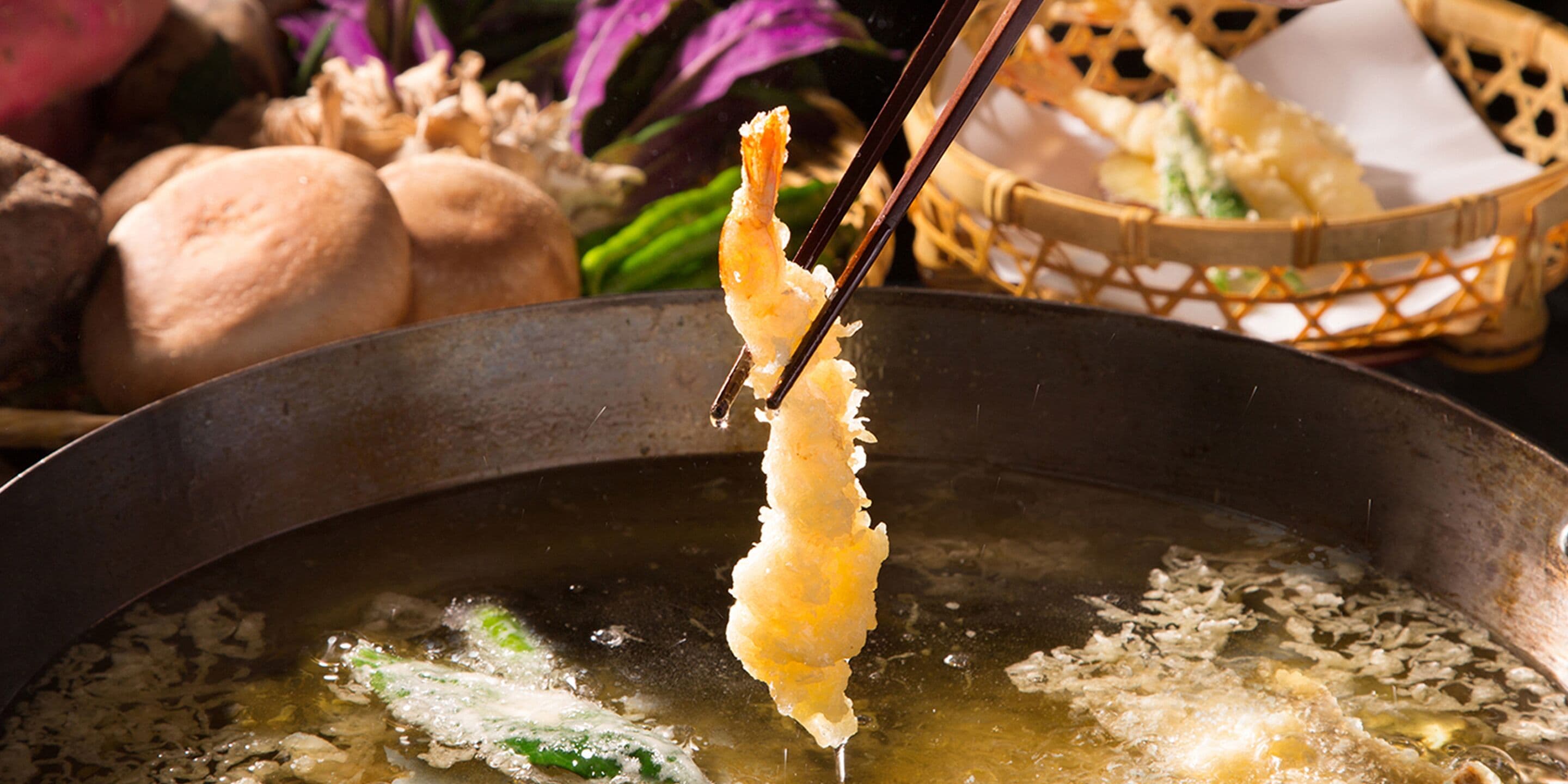 A close-up of a food item being held with chopsticks over a pot of boiling liquid, with a basket of food items in the background.