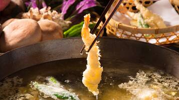 A close-up of a food item being held with chopsticks over a pot of boiling liquid, with a basket of food items in the background.