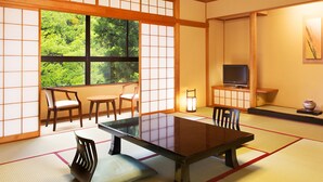 Una habitación japonesa tradicional con suelo de tatami, una mesita baja, sillas, un televisor y vistas a zonas verdes a través de un ventanal grande.