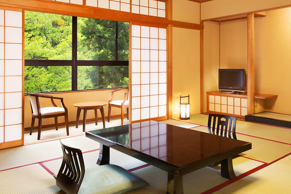 Habitació japonesa tradicional amb sòls de tatami, una taula baixa, cadires, televisió i vista a la vegetació a través d'una finestra gran.