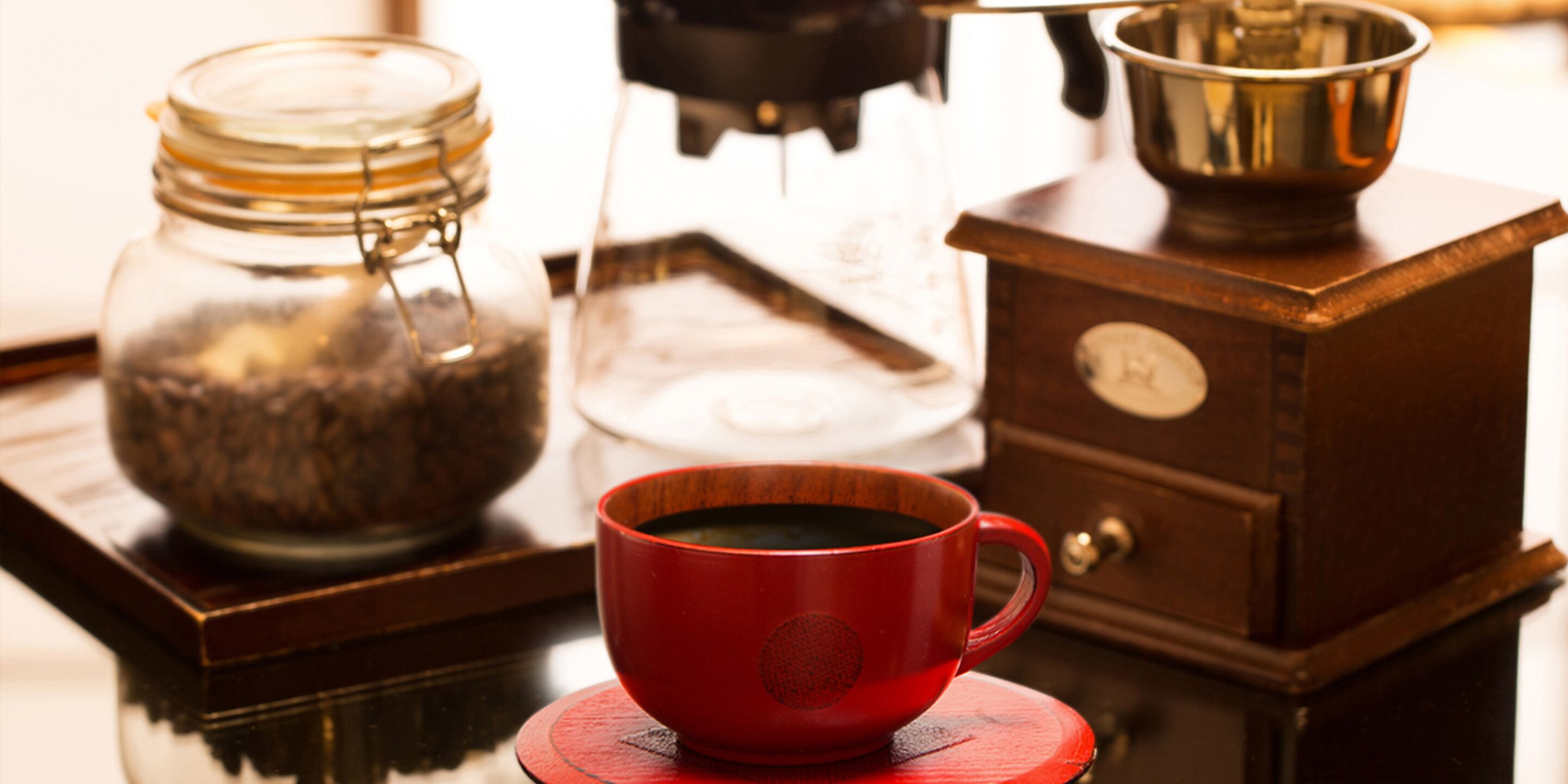 A coffee maker, a red cup, and a glass jar on a wooden table.